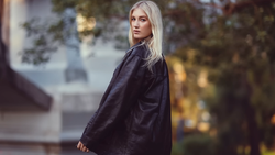 Woman with long platinum blonde hair wearing a dark navy jacket, photographed outdoors in soft golden-hour light.