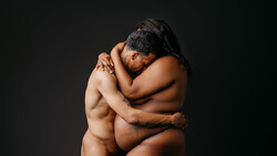 Two shirtless men embracing against a dark background, shot in studio lighting.