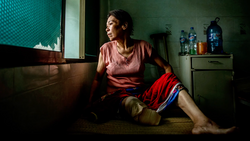 Woman in pink shirt sitting on floor in modest room, gazing toward window with natural light.