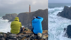 Two photographers in bright jackets observe a dramatic rocky coastline with a red and white lighthouse and turbulent ocean waves.