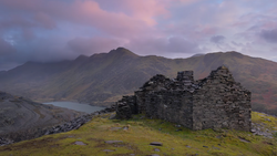 Ruined stone cottage on grassy hillside with mountains and water under dramatic twilight sky.