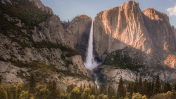 Tall waterfall cascading down a granite cliff face between massive rock formations at golden hour.