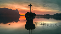Silhouetted crosses on a rocky outcrop in still water at sunrise, with mountains and forested shorelines in the background.