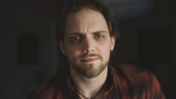 Man with dark hair and beard wearing a red plaid shirt, looking directly at camera with a serious expression.