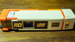 Lomo Matic 110 instant film camera with orange and white body on wooden surface.