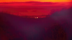 Silhouetted mountain ridges against a deep red and orange sunset sky with two distant lights visible on the horizon.