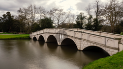 Stone arch bridge spanning a calm river with bare trees and green lawn in overcast daylight.