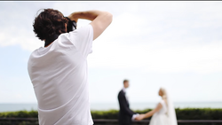 Photographer in white shirt capturing a wedding couple with camera held to face, bride and groom visible in soft-focus background.