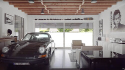 Black Porsche 911 displayed in a minimalist white garage with exposed wooden ceiling beams.