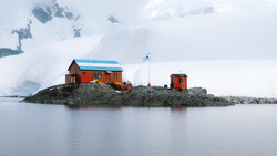 Orange and red buildings with blue roof on rocky island surrounded by icy water and snow-covered mountains.