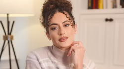 Woman with curly dark hair styled in a half-up bun, wearing a light pink ribbed sweater, posing with hand near face in a bright interior setting.