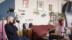 Filmmaker operating camera on tripod while interviewing subject seated on burgundy couch in home studio.