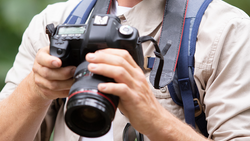 Photographer holding a DSLR camera with telephoto lens, wearing a camera strap and light-colored shirt.