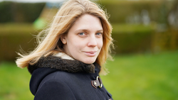 Woman with blonde hair looking over shoulder outdoors, wearing black jacket with fur collar.
