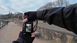 Photographer's hands holding a smartphone to frame a riverside promenade with bare trees and stone walls.