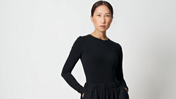 Woman in black long-sleeve top and gathered skirt standing against neutral background.