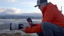 Photographer holding a telephoto lens with red hood near a frozen lake, mountains visible across water.