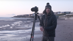 Photographer with camera on tripod on a beach at dusk, wearing winter clothing and beanie.