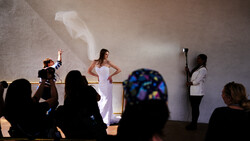 Woman in white dress posing on stage during a fashion or bridal show while photographers and spectators watch from the audience.