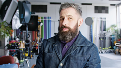 Bearded man in denim jacket inside a photography studio with organized equipment and lighting setups visible in the background.