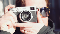 Person holding a rangefinder film camera with a fixed lens, wearing tortoiseshell sunglasses.