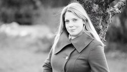 Woman with long blonde hair wearing a collared coat, positioned against a tree in an outdoor setting.