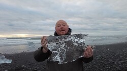 Man holding a large chunk of ice on a black sand beach with ocean and cloudy sky behind him.