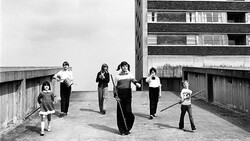 Children playing with a large tire on a rooftop concrete platform beside a brutalist apartment building.