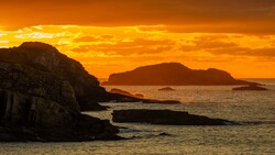 Silhouetted rocky coastline and islands against a golden orange sunset sky.