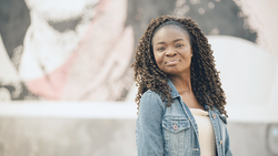 Woman with long braided hair wearing a denim shirt smiles at camera against blurred urban background.
