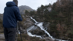 Photographer on tripod capturing a cascading mountain stream surrounded by evergreen forest and rocky terrain.
