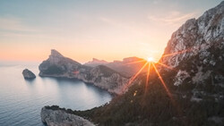 Sun bursting through rocky cliffs above a calm Mediterranean cove at sunrise.