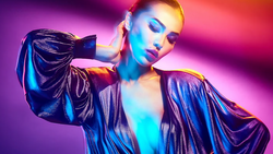 Person with short blonde hair wearing iridescent jacket against vibrant gradient background with blue and pink lighting.