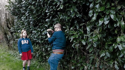Photographer taking a portrait of a young girl standing against dense ivy-covered foliage.