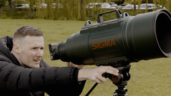 Photographer holding a large black telephoto lens marked with orange Sigma branding outdoors.