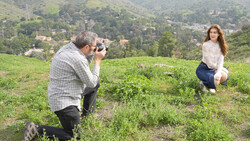 Photographer crouching while shooting a model posing in a grassy hillside field with forested mountains in the background.