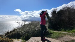 Person in magenta jacket photographing a coastal landscape with binoculars or telephoto lens.