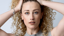Woman with voluminous curly blonde hair posing with hands near head against neutral background.