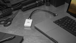 Creative professional's desk setup with laptop, external hard drive, camera equipment, and accessories arranged on gray surface.