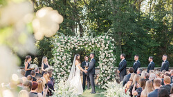 Bride and groom exchanging vows beneath an elaborate floral arch at an outdoor wedding ceremony.