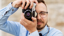 Photographer wearing glasses looking through viewfinder of rangefinder camera in light blue shirt.