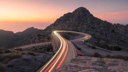 Long exposure light trails curve through a desert mountain pass at sunset.
