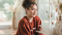 Woman in rust-colored sweater gazing to the side near a sunlit window.