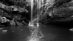 Waterfall cascading into a rocky pool surrounded by layered stone formations in black and white.