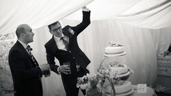 Two men in formal attire gesture toward a tiered wedding cake in a white draped tent setting.