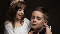 Young girl with tiara embracing boy from behind against dark background.