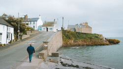 Person jogging along a coastal road with stone buildings and cliff edge visible.
