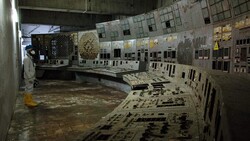 Abandoned control room with operator station and wall-mounted instrument panels covered in dust and decay.