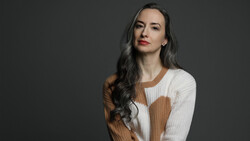 Woman with long dark hair wearing a cream and tan ribbed knit sweater against a gray background.