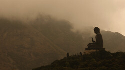Large Buddha statue silhouetted against misty mountains shrouded in fog and clouds.